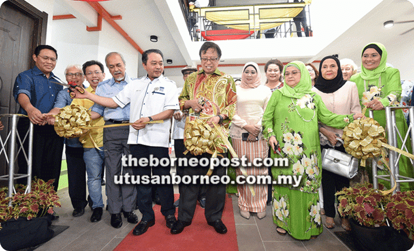 Group in colorful attire at indoor ribbon-cutting ceremony, smiling with golden ribbon bows, conveying celebration.