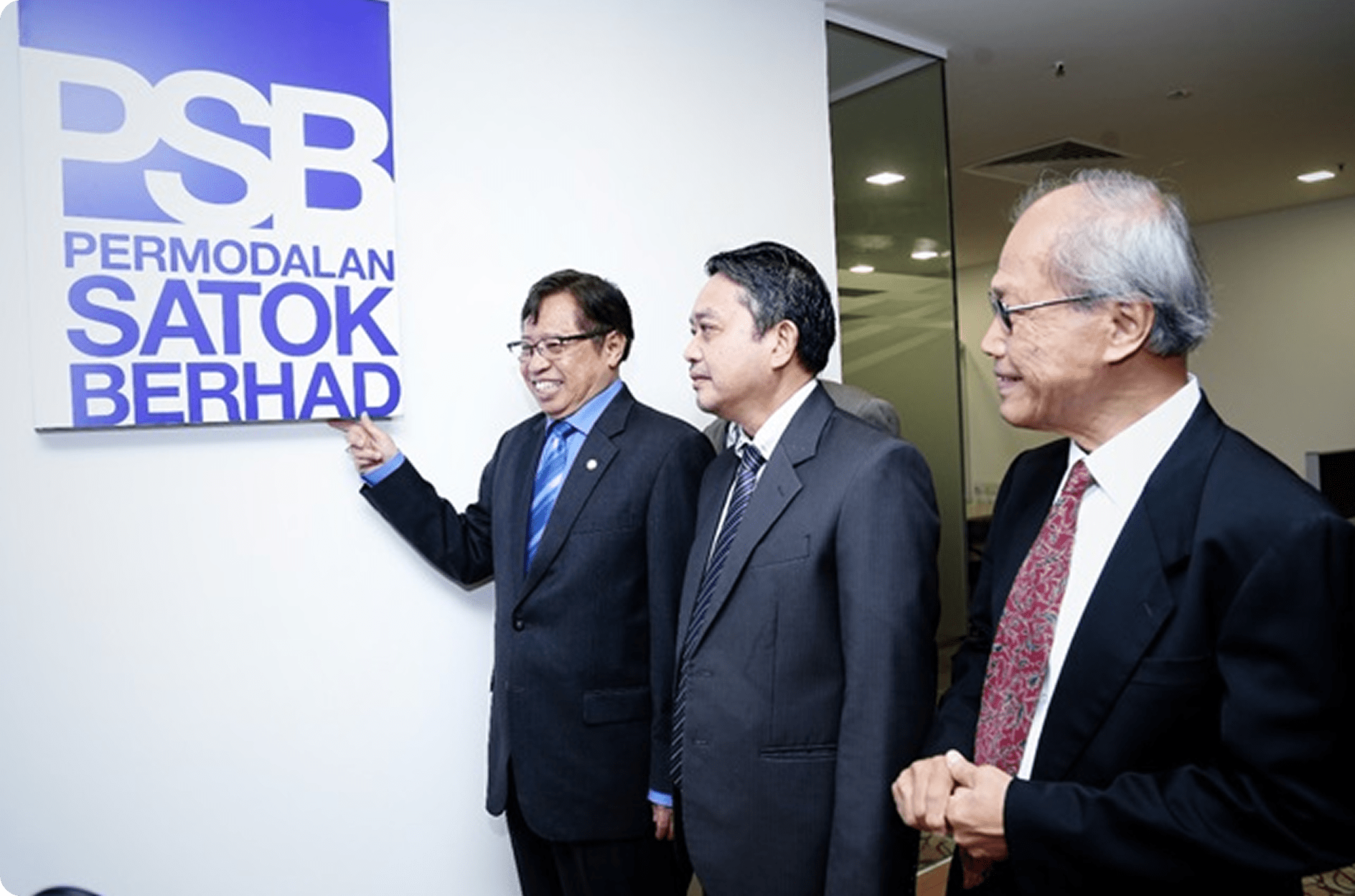 Three suited men smile beside a "PSB Permodalan Satok Berhad" sign in a bright office, conveying professionalism.