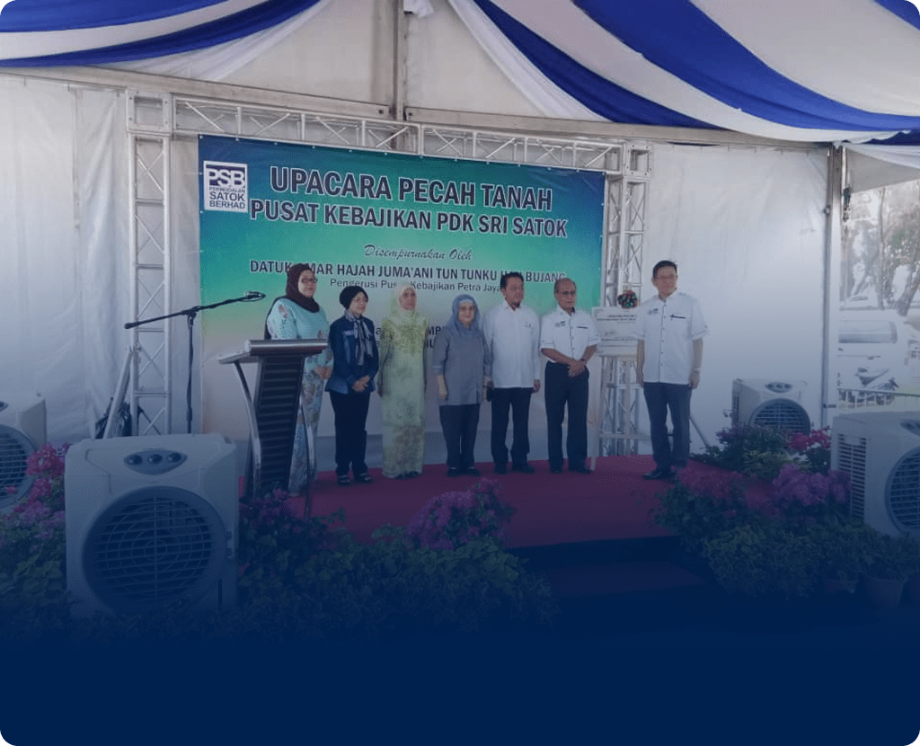 Group on stage under white-blue tent at Pusat Kebajikan PDK Sri Satok groundbreaking, with event sign behind.
