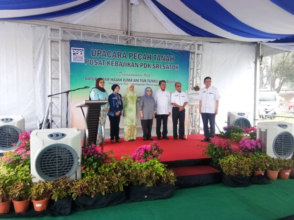 Seven people on stage at a welfare center groundbreaking under a blue-white tent. Formal atmosphere.