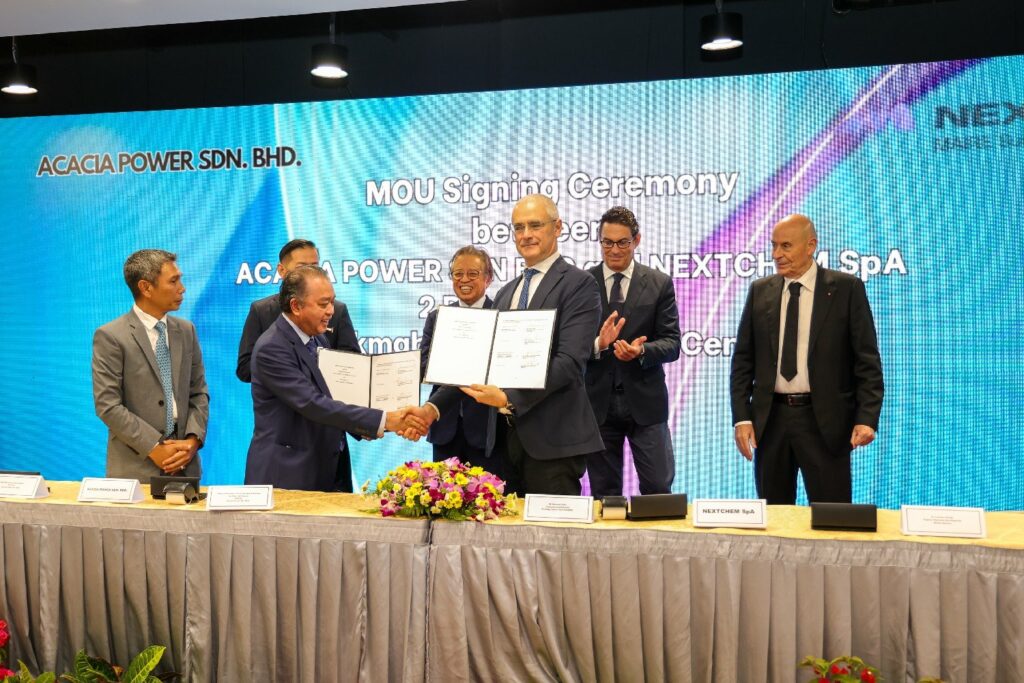 Seven men stand before an "MOU Signing" screen; two exchange documents with a handshake, marking a formal occasion.