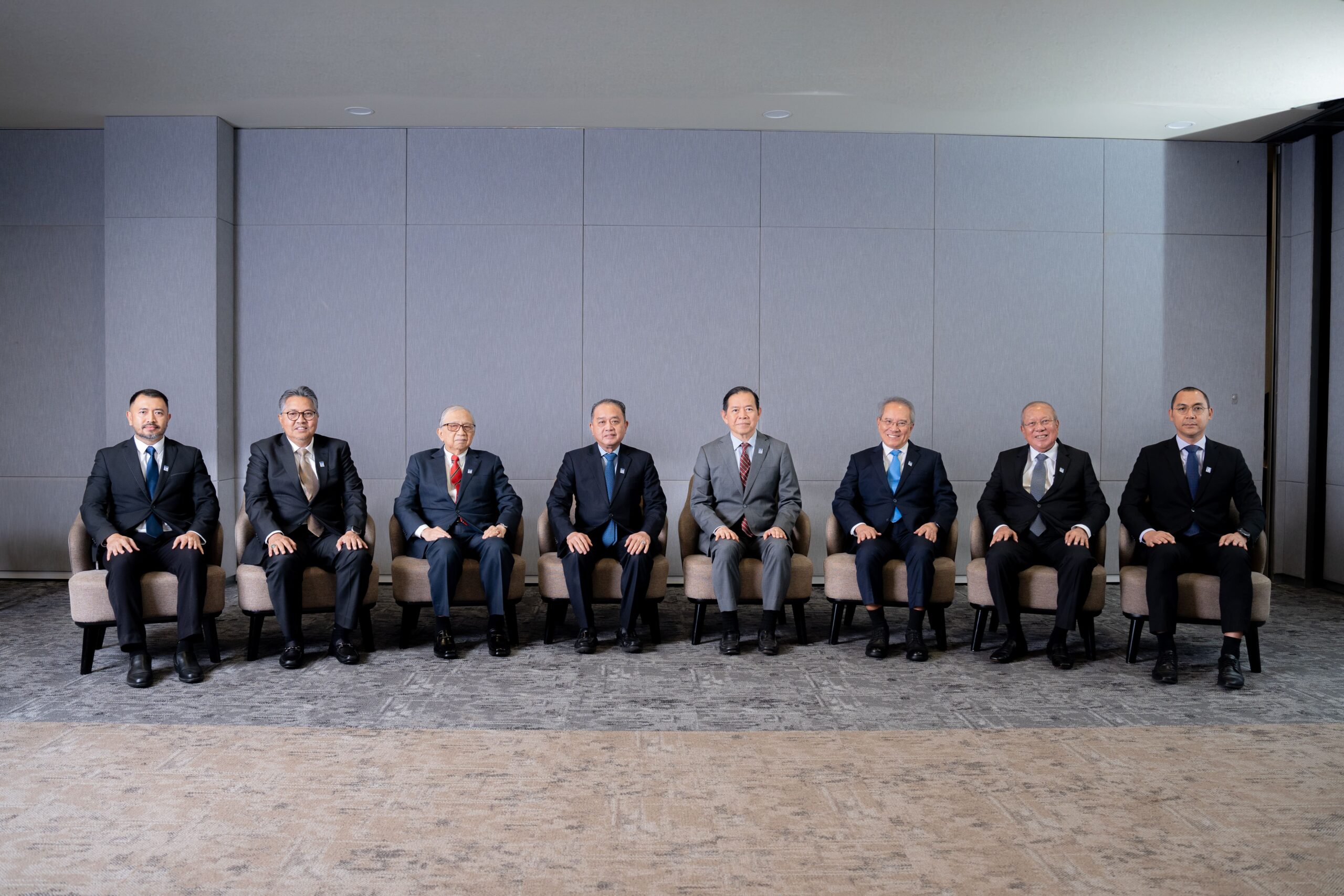 Eight men in suits sit in a row against a gray wall, in a spacious, well-lit room, projecting a formal and professional vibe.
